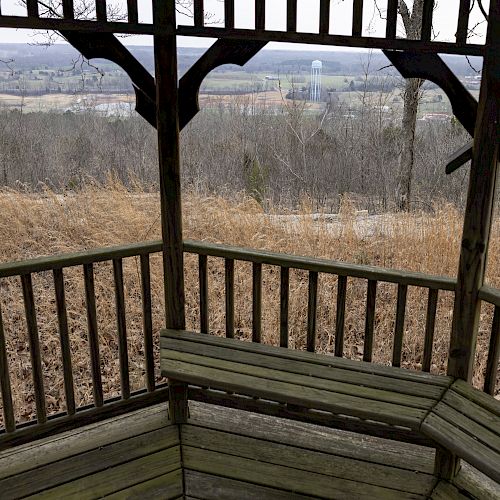 A wooden gazebo overlooking a scenic, rural landscape with dry grass and bare trees, and distant hills and structures under a cloudy sky.