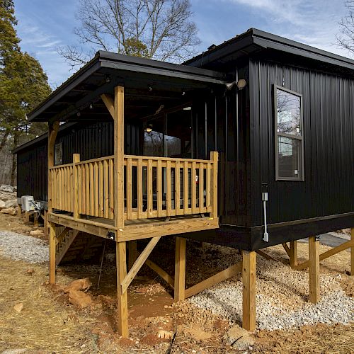 A small black cabin on stilts with a wooden deck and railing, set in a wooded area with bare trees and a dirt and gravel path.