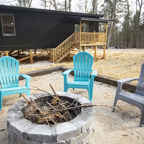 The image shows a fire pit surrounded by two blue Adirondack chairs and one gray chair, with a black cabin featuring a porch in the background.