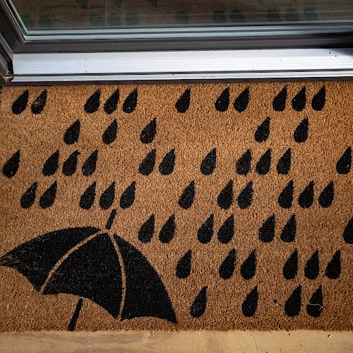 Doormat on wooden floorboards, featuring black raindrop patterns with an umbrella illustration in the lower left corner.