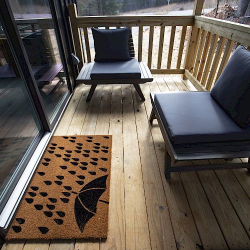 A cozy wooden deck with two cushioned chairs and a doormat featuring an umbrella design with raindrops near a sliding glass door.