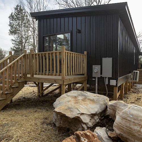 A modern black tiny house with a wooden porch and steps, situated in a rural area with large rocks and trees surrounding the property.