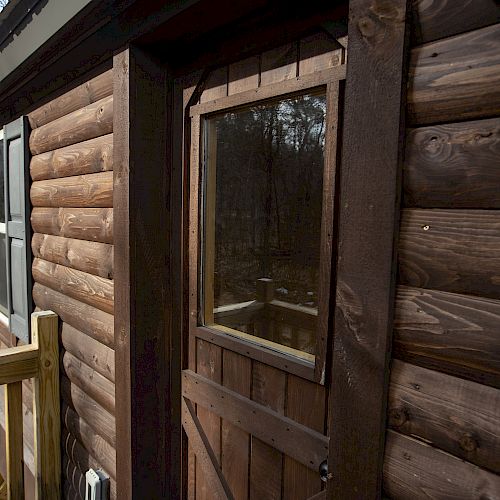 This image shows a rustic wooden cabin with a door and a window, situated in a wooded area with a wooden deck railing visible.