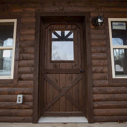 A wooden cabin door is flanked by two windows, with a light fixture above and an electrical box to the left of the door.