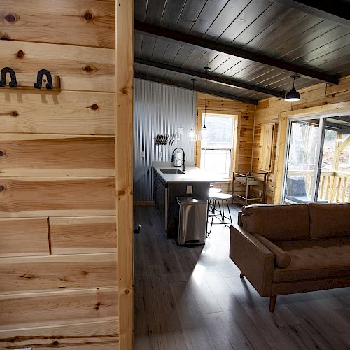 A cozy, modern cabin interior with wooden walls, a couch, a kitchen area, and large windows letting in natural light, creating a warm atmosphere.