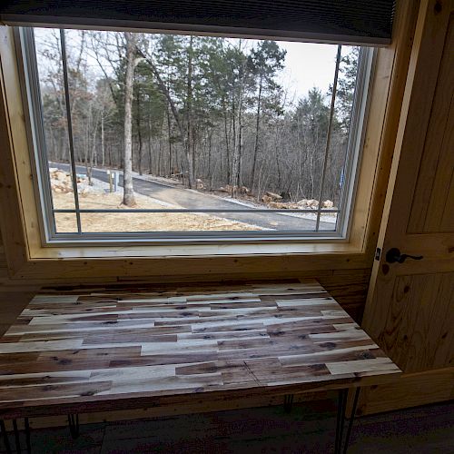 The image shows a wooden table in front of a window with a view of a forest and a road outside. The interior is wood-paneled, including the door.