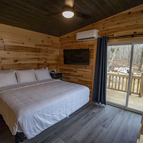 A cozy cabin room with a large bed, wooden walls, a mounted TV, an air conditioner, and a sliding glass door leading to a deck with an outdoor view.