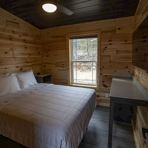 A cozy wooden cabin bedroom with a bed, bedside tables, a window, and a wall-mounted TV. The room features warm wood paneling.