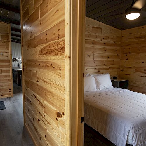 A cozy, wooden cabin interior featuring a hallway leading to a bedroom with a made bed and window, and a partially visible bathroom beyond the hallway.