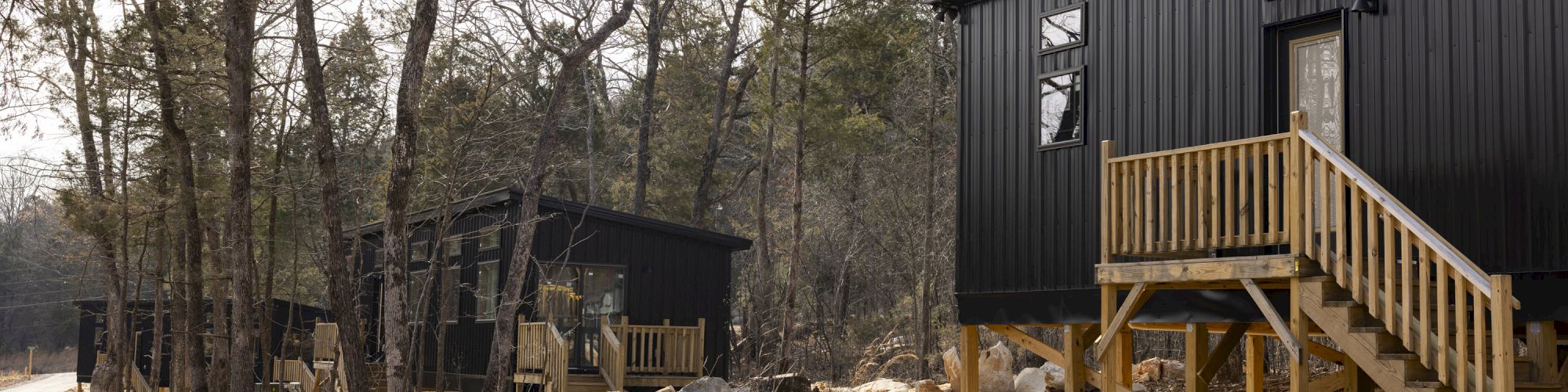 The image shows black tiny houses in a wooded area with elevated wooden decks and stairs, surrounded by leafless trees on a sunny day.