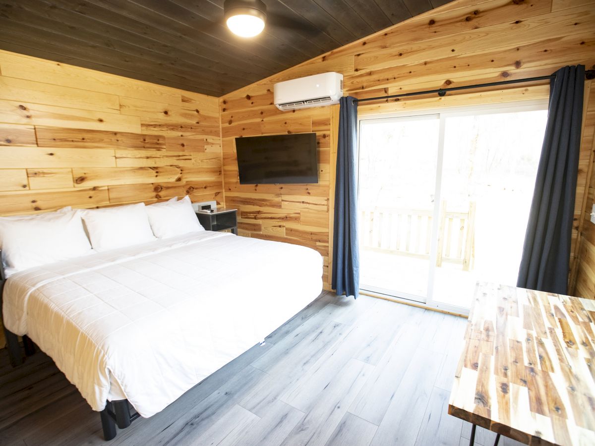 A cozy wooden bedroom with a large bed, wall-mounted TV, small desk, and sliding glass door with curtains leading to a balcony.