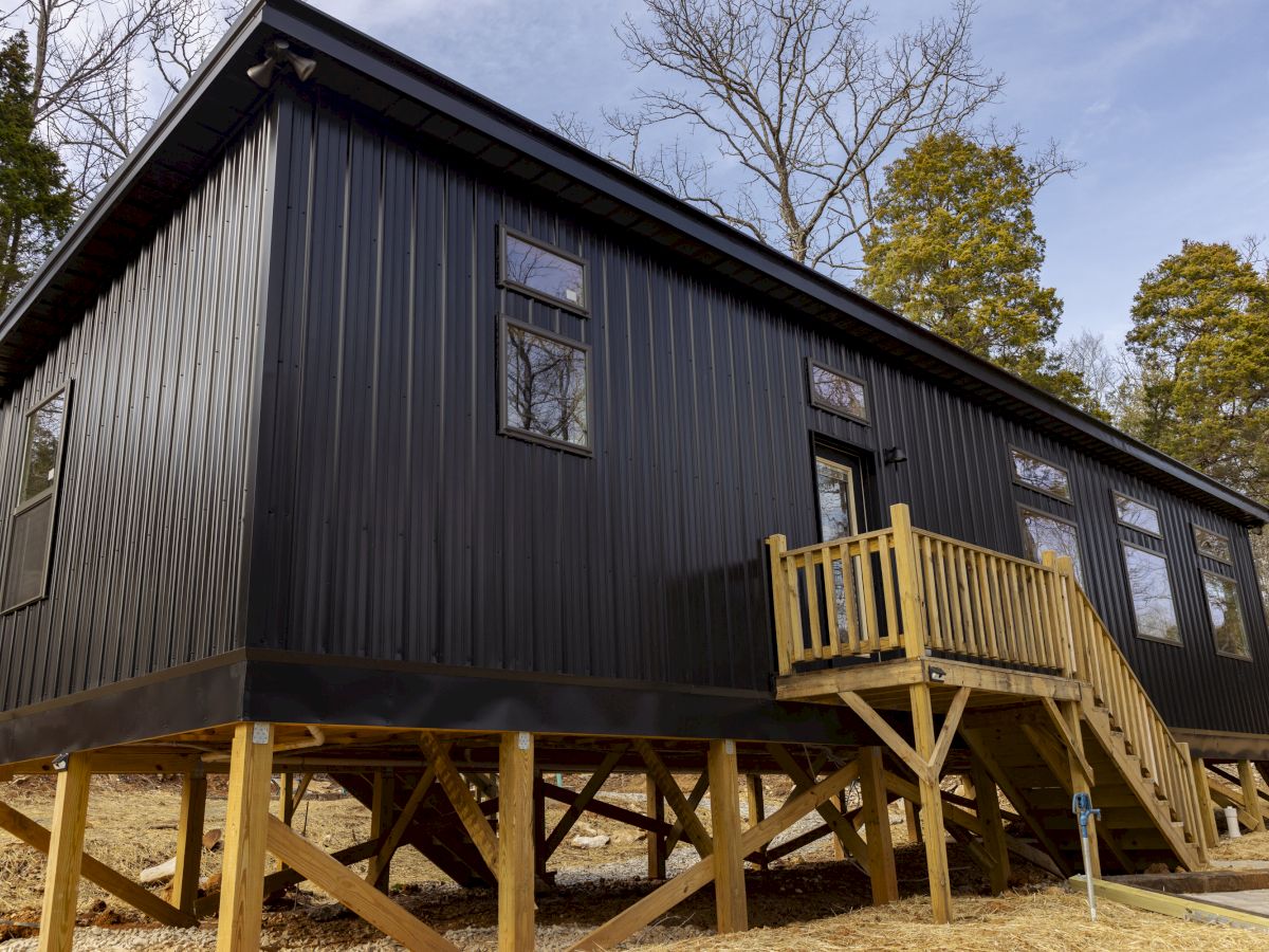 The image shows a black elevated cabin with a metal exterior, wooden stairs, and multiple windows, set against a backdrop of trees in a natural area.