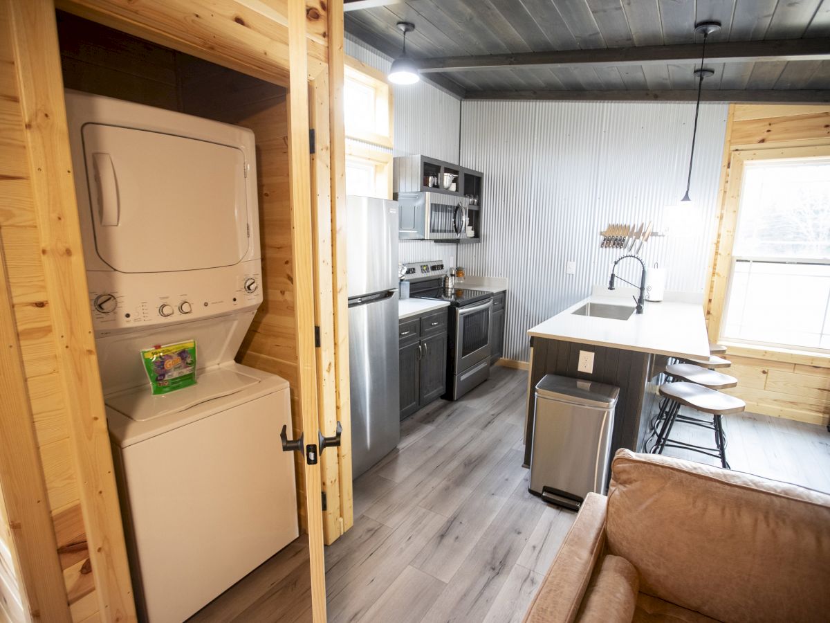 A cozy kitchen and laundry area featuring wooden accents, a stacked washer-dryer, stainless steel appliances, and light streaming through the windows.