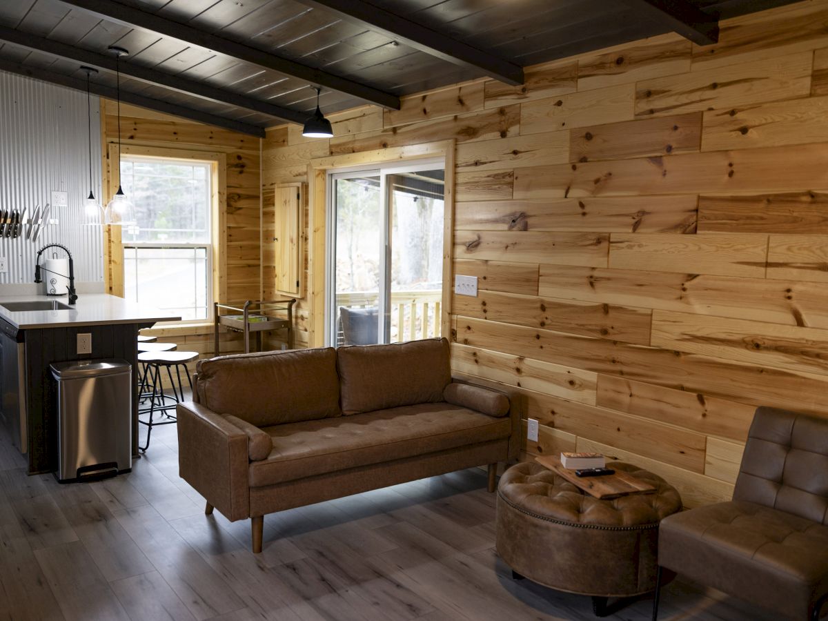 A cozy living area with wood panel walls, featuring a couch, round ottoman, chair, open kitchen, and dining area with natural light.