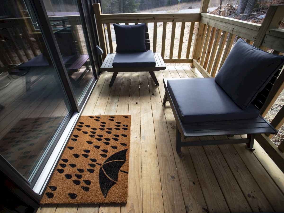 A small wooden porch with two cushioned chairs and a doormat featuring an umbrella design with raindrops, next to a sliding glass door.