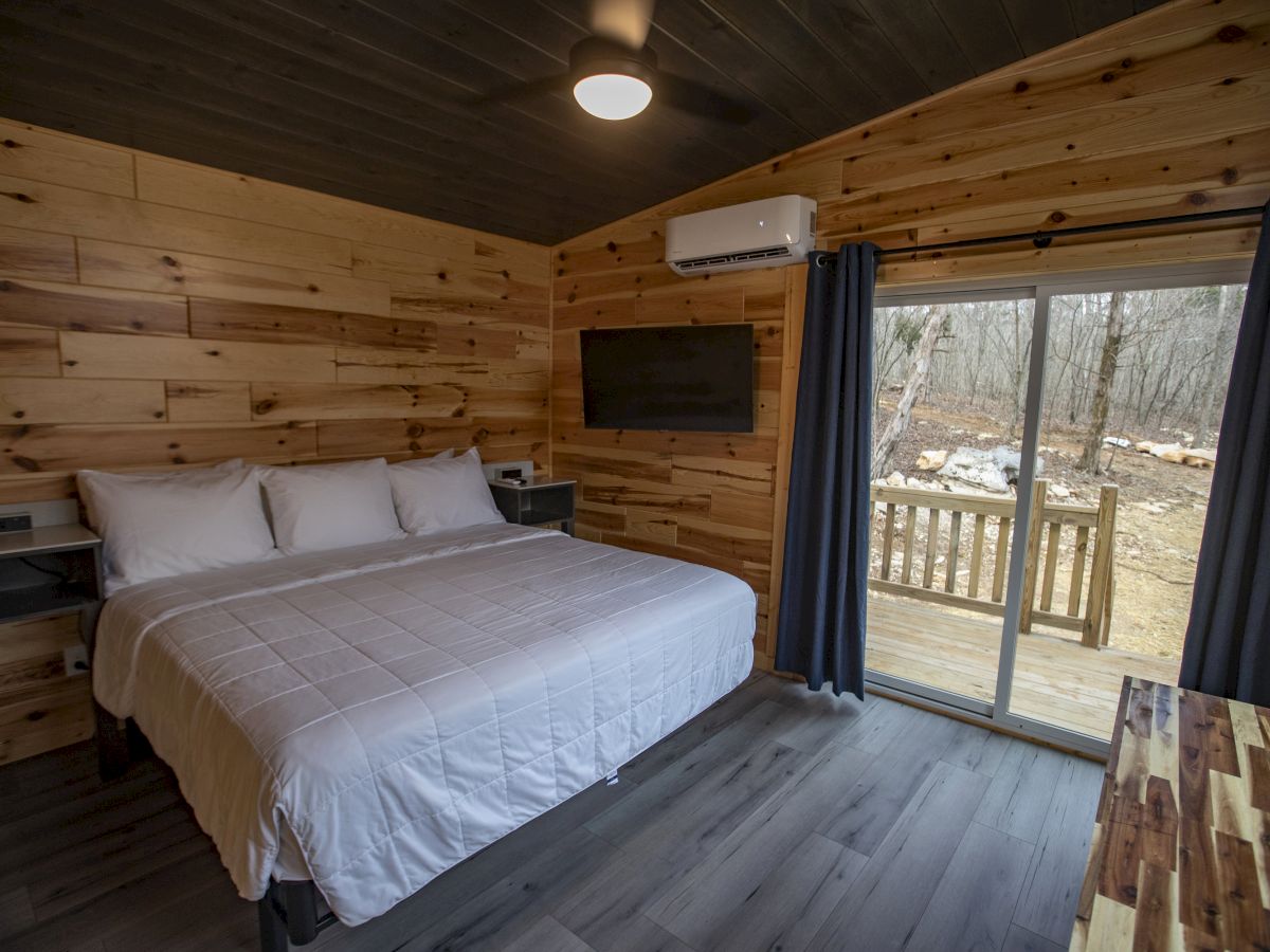 A cozy cabin room with a large bed, wood paneling, a wall-mounted TV, sliding glass door with curtains, and a view of the outdoors.