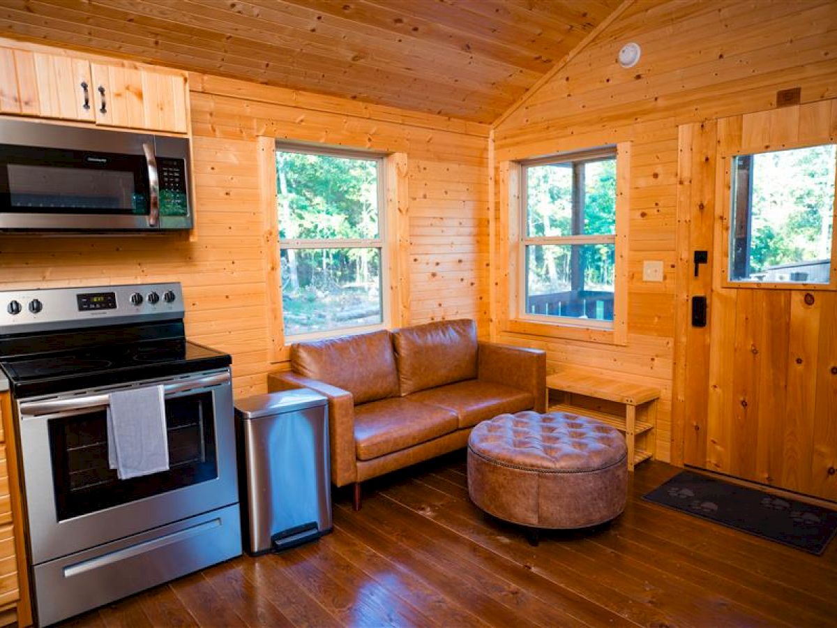 A cozy wooden cabin interior with a stove, microwave, leather couch, round ottoman, and two windows near a door, all made of wood.