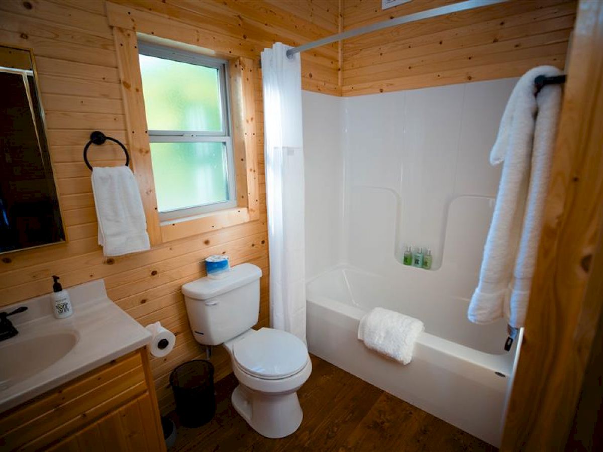 A cozy wooden bathroom with a bathtub, shower, toilet, sink, and mirror. Towels are neatly hung, and there is a small window for natural light.