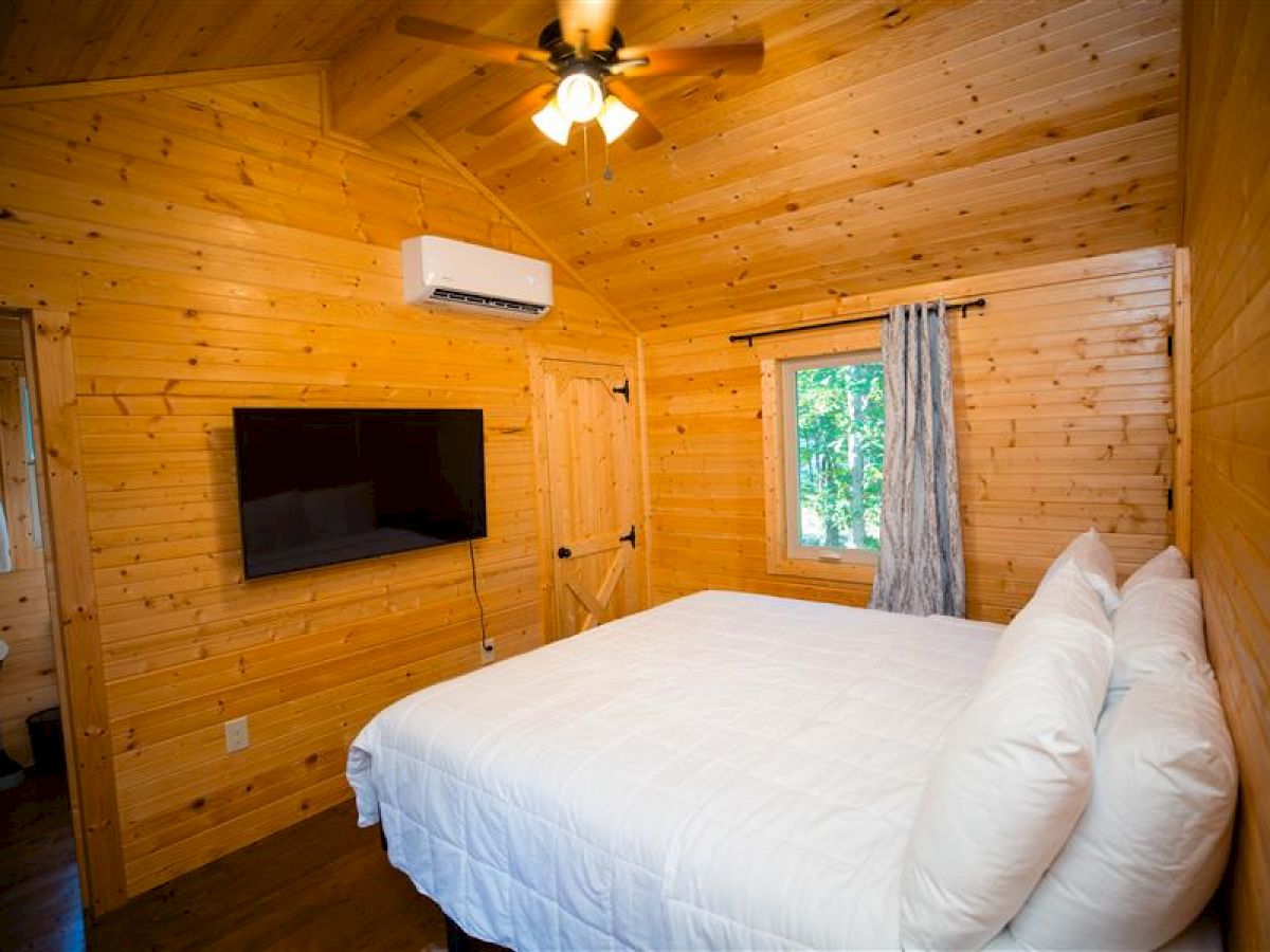 A cozy wooden cabin bedroom with a double bed, a wall-mounted TV, a ceiling fan, a window with curtains, and an air conditioner.