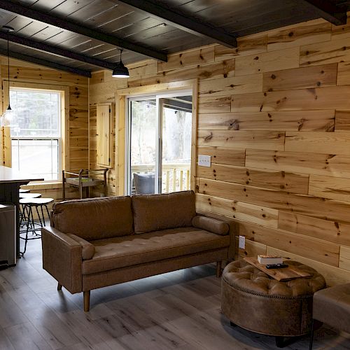 A cozy living space with wooden walls, a brown couch, a round ottoman, an armchair, and an open kitchen in the background.