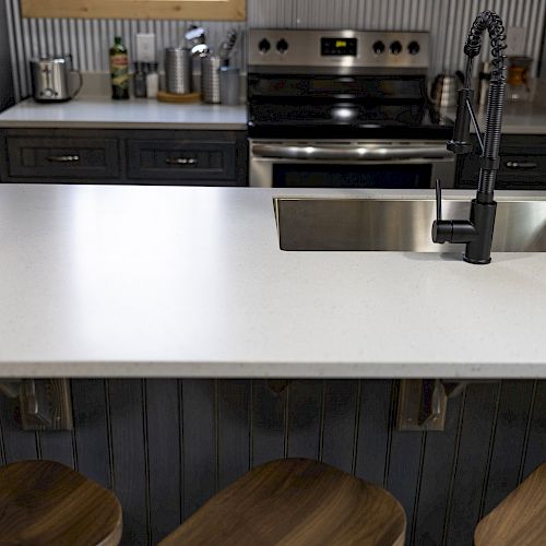 A modern kitchen features a white countertop island with a sink and black faucet, accompanied by wooden stools, stainless steel appliances in the background.