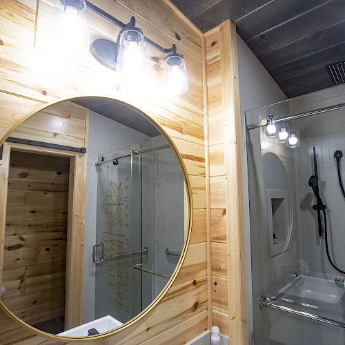 This image shows a modern bathroom with a circular mirror, wooden walls, shelf lights, and a glass shower enclosure with a rainfall showerhead.