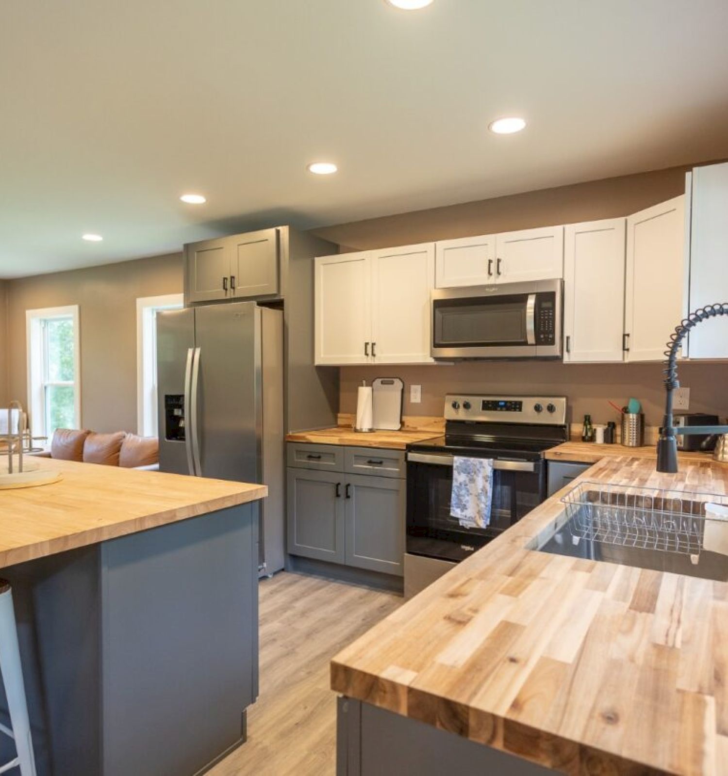 A modern kitchen with wooden countertops, stainless steel appliances, white cabinets, and bar stools at the island, connects to a living area.
