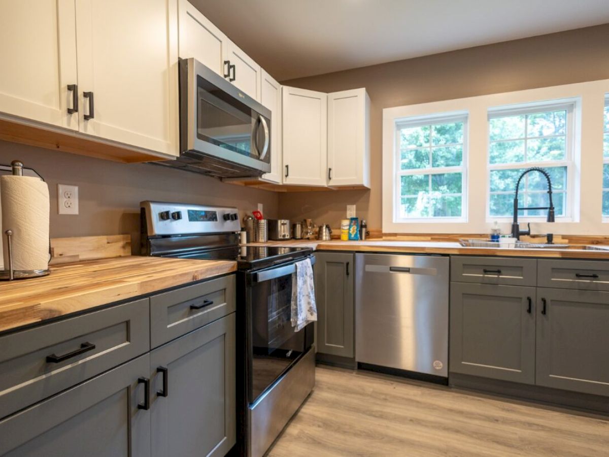 A modern kitchen with two-tone cabinets, stainless steel appliances, and a paper towel holder, under natural light from three windows.