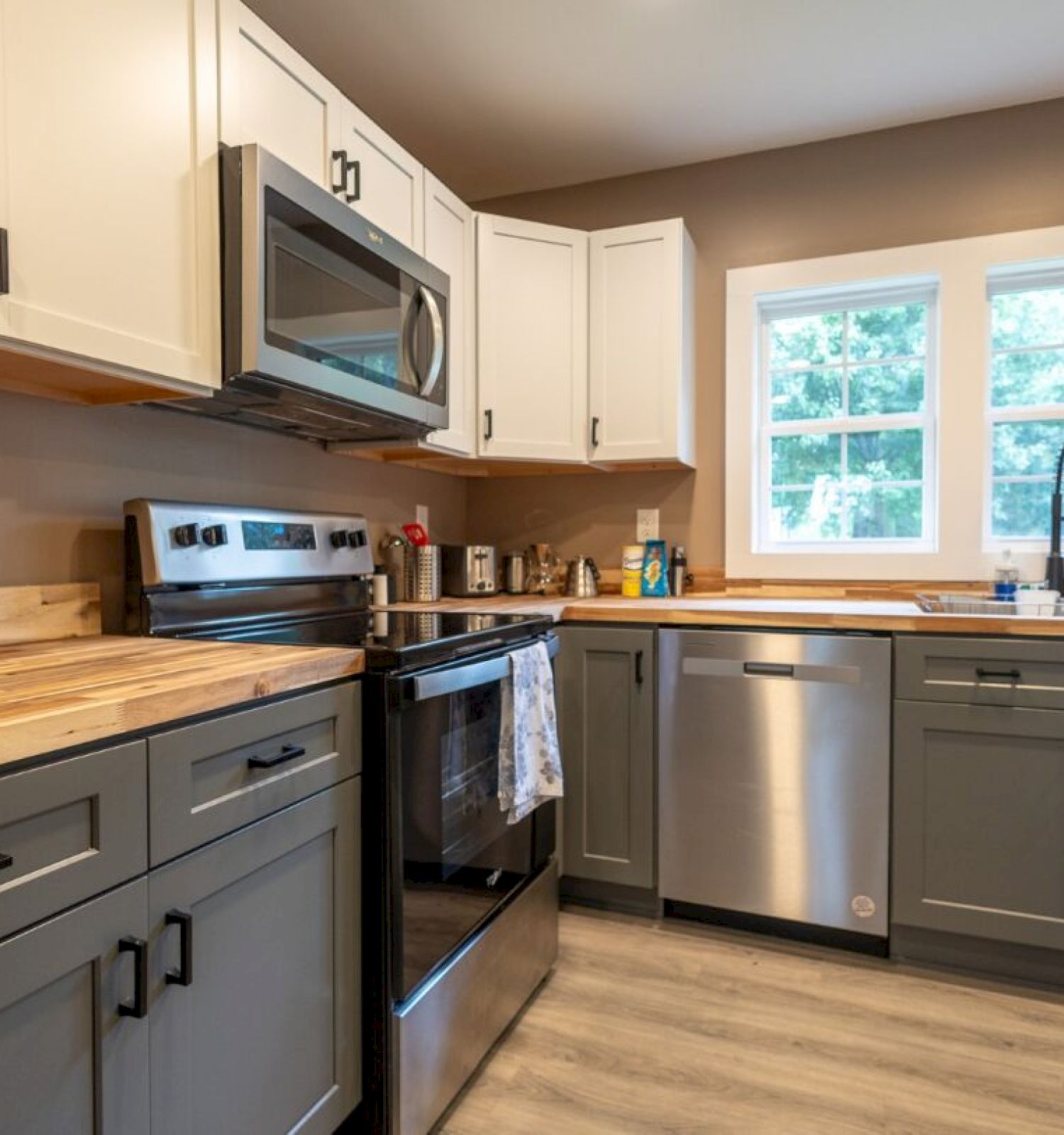 A modern kitchen with two-tone cabinets, stainless steel appliances, and a paper towel holder, under natural light from three windows.