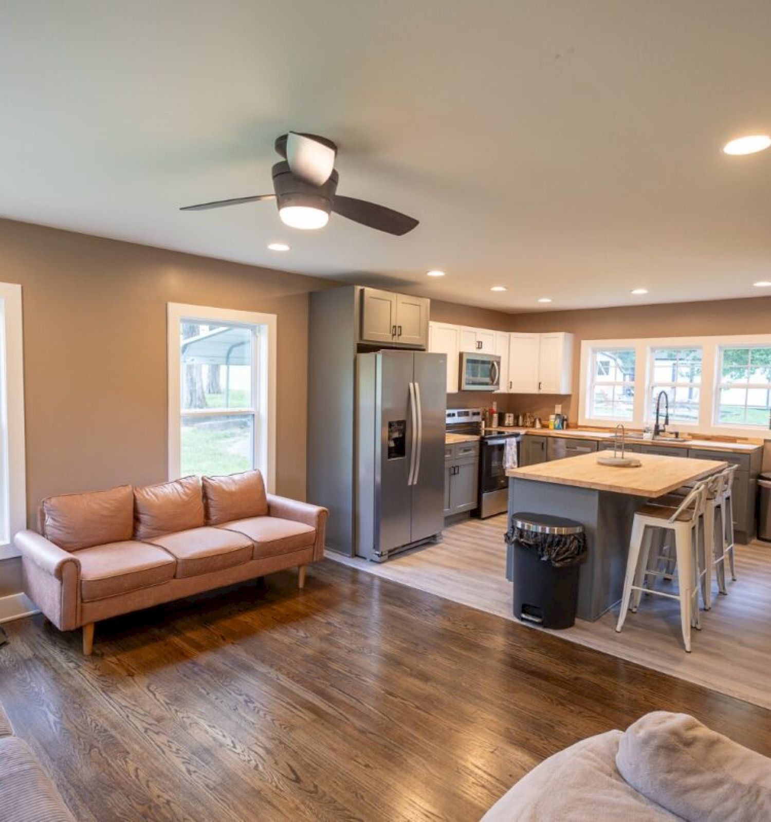 A cozy open-concept living room and kitchen with hardwood floors, sofas, a ceiling fan, stainless steel appliances, and an island.