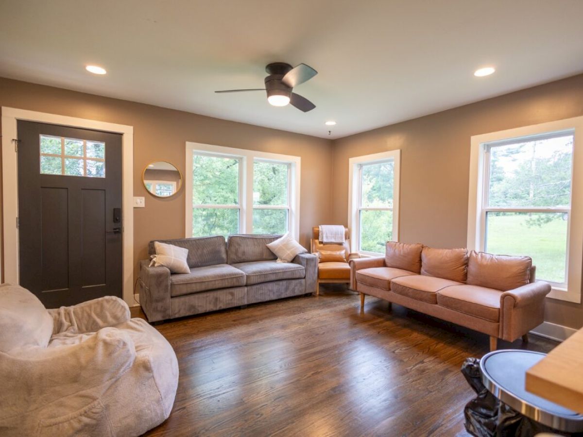 A cozy living room with two sofas, an armchair, a dark door, wooden floors, windows, a ceiling fan, and soft lighting.