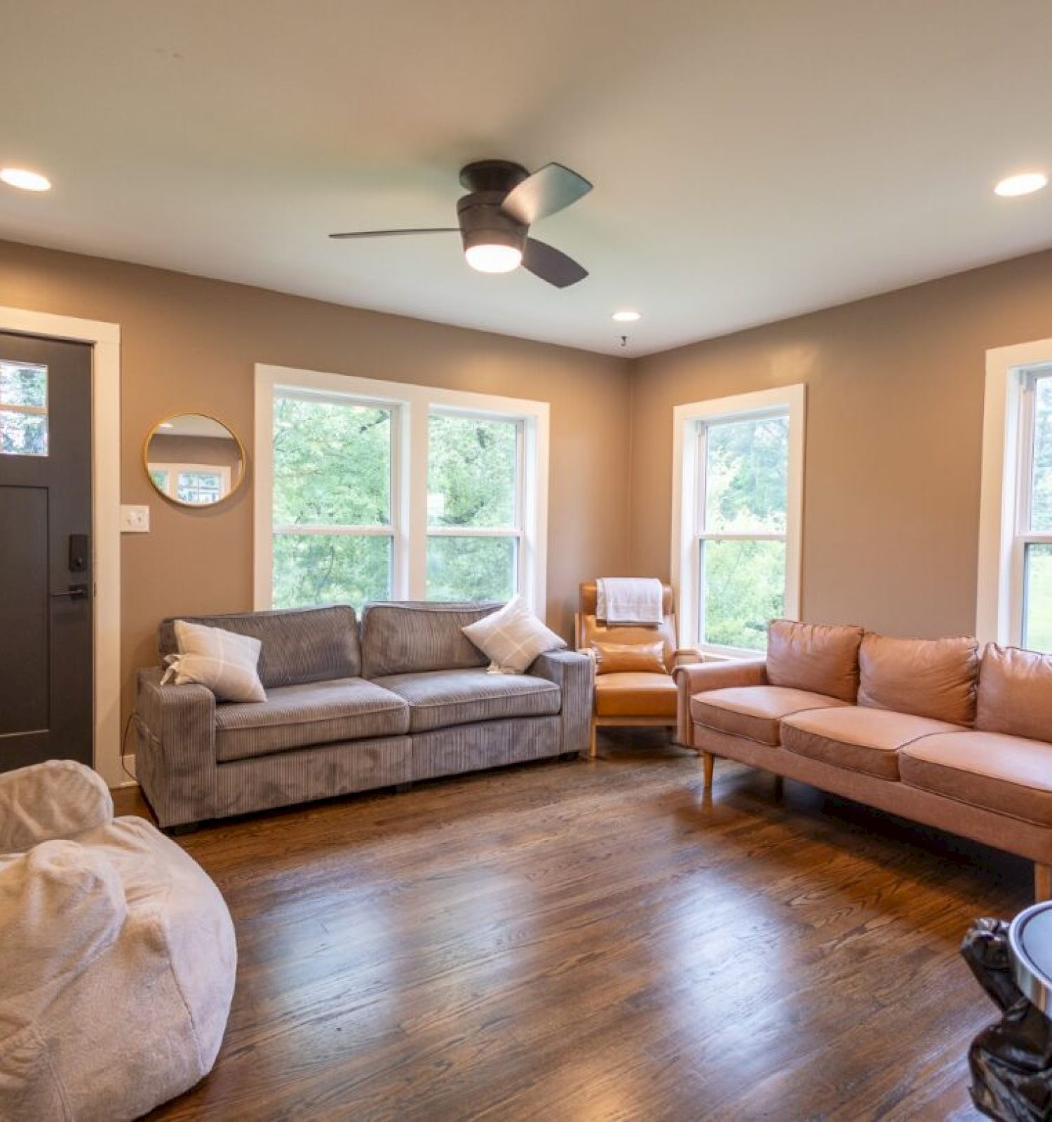 A cozy living room with two sofas, an armchair, a dark door, wooden floors, windows, a ceiling fan, and soft lighting.