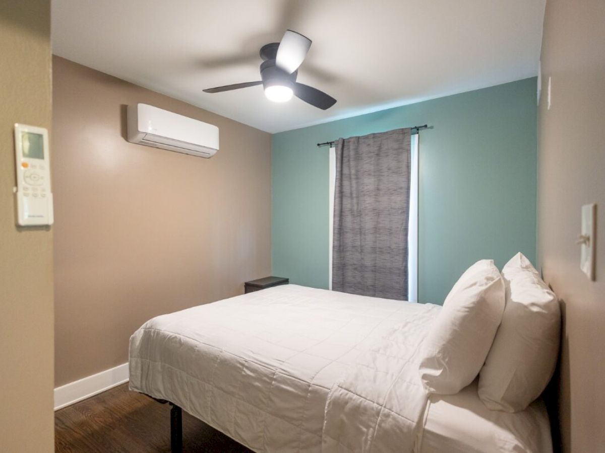 A bedroom with a bed, white bedding, a ceiling fan, and a wall-mounted air conditioner. There's a window with a grey curtain.