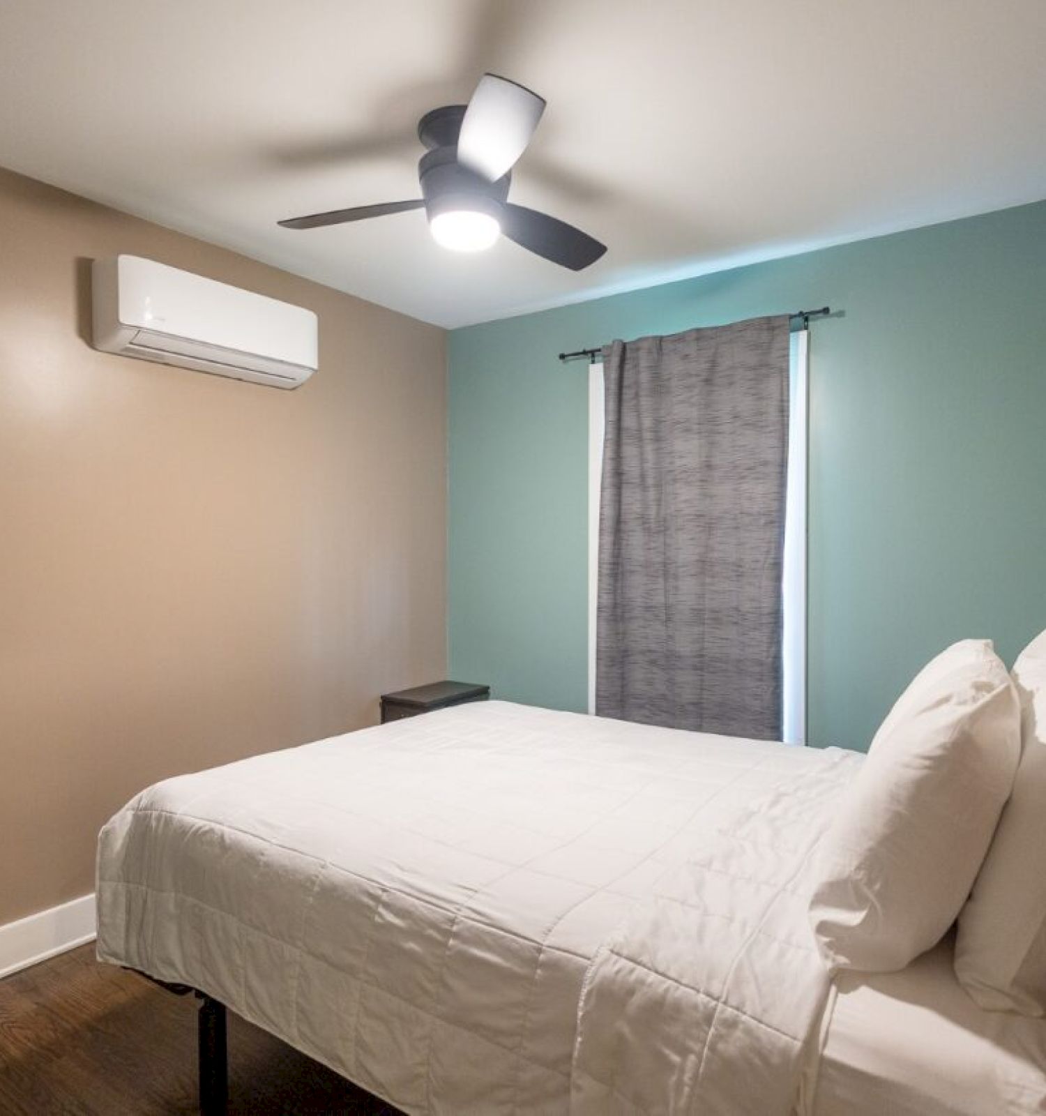 A bedroom with a bed, white bedding, a ceiling fan, and a wall-mounted air conditioner. There's a window with a grey curtain.