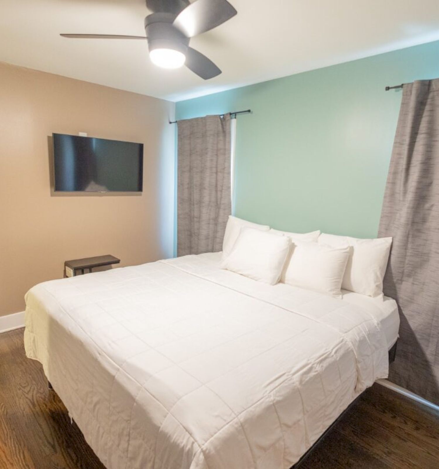 The image shows a bedroom with a large bed, a ceiling fan, a wall-mounted TV, and curtains.