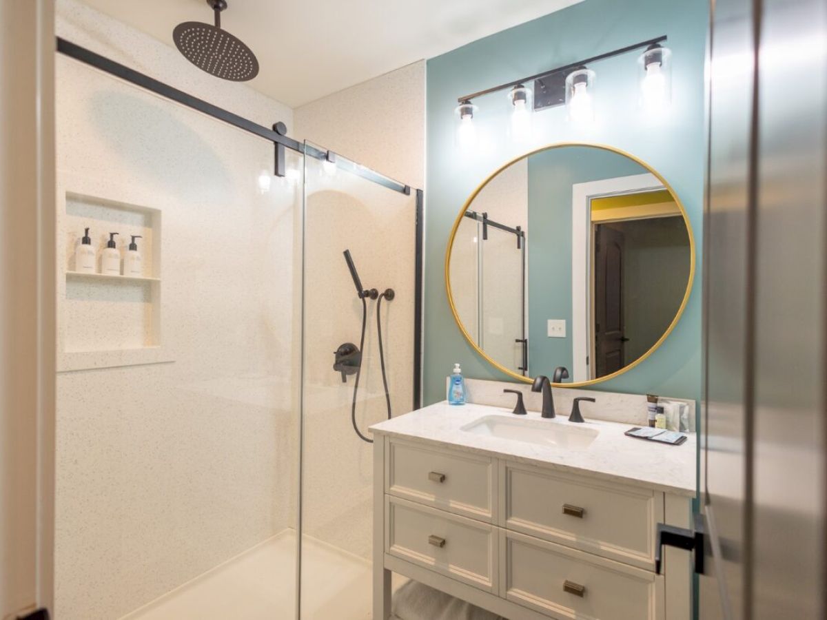 The image shows a modern bathroom with a glass shower, vanity with round mirror, and sleek lighting above.