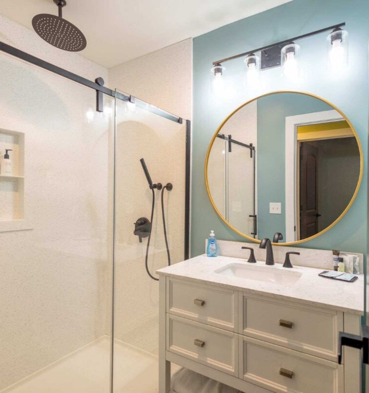 The image shows a modern bathroom with a glass shower, vanity with round mirror, and sleek lighting above.