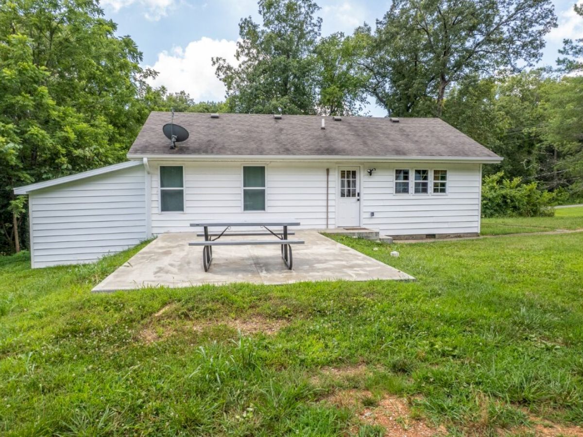 A small white house with a gabled roof, a satellite dish, a patio with a picnic table, and surrounding trees and grass are featured in the image.