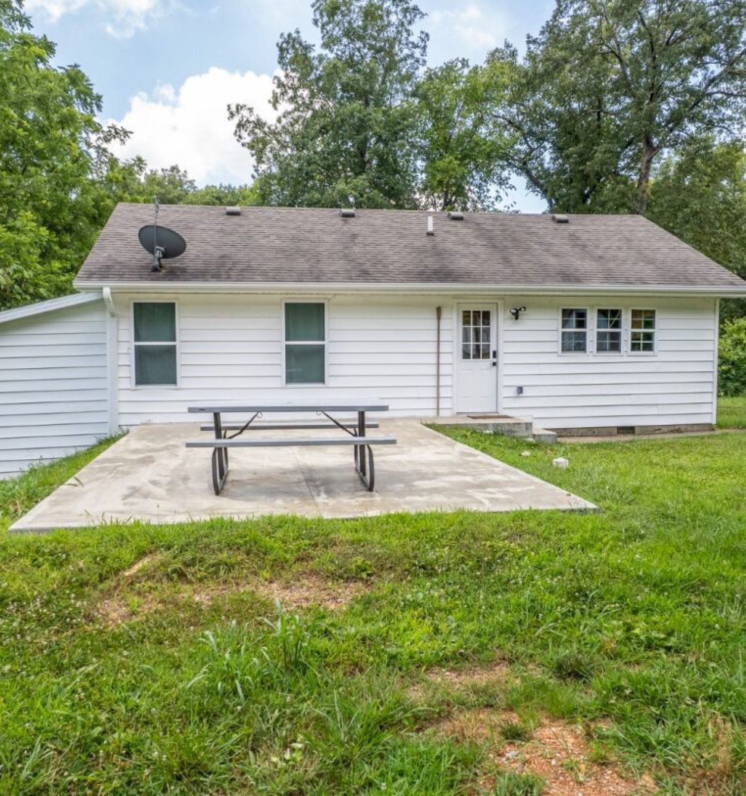 A small white house with a gabled roof, a satellite dish, a patio with a picnic table, and surrounding trees and grass are featured in the image.