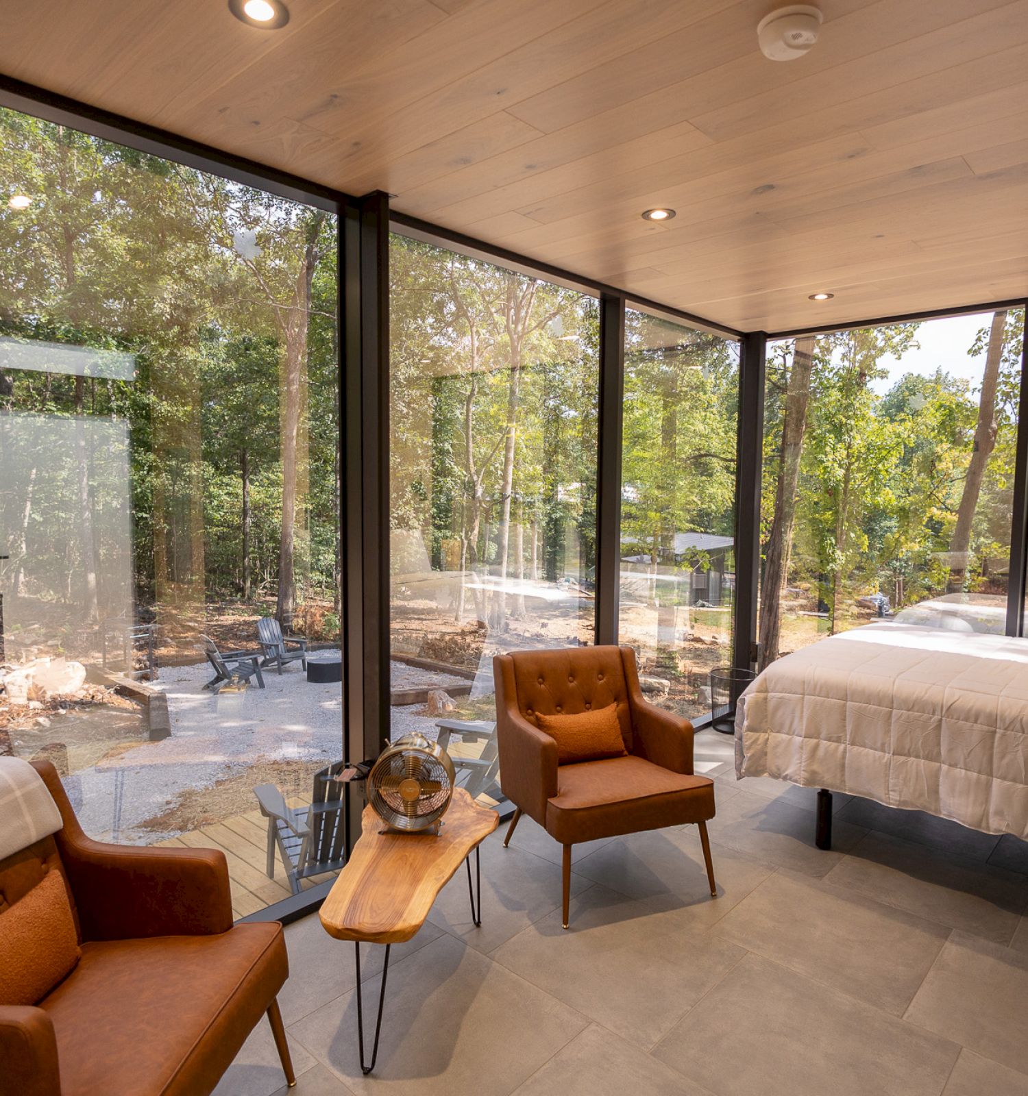 A cozy room with large windows, two armchairs, a small table, and a bed, overlooking a forested view.