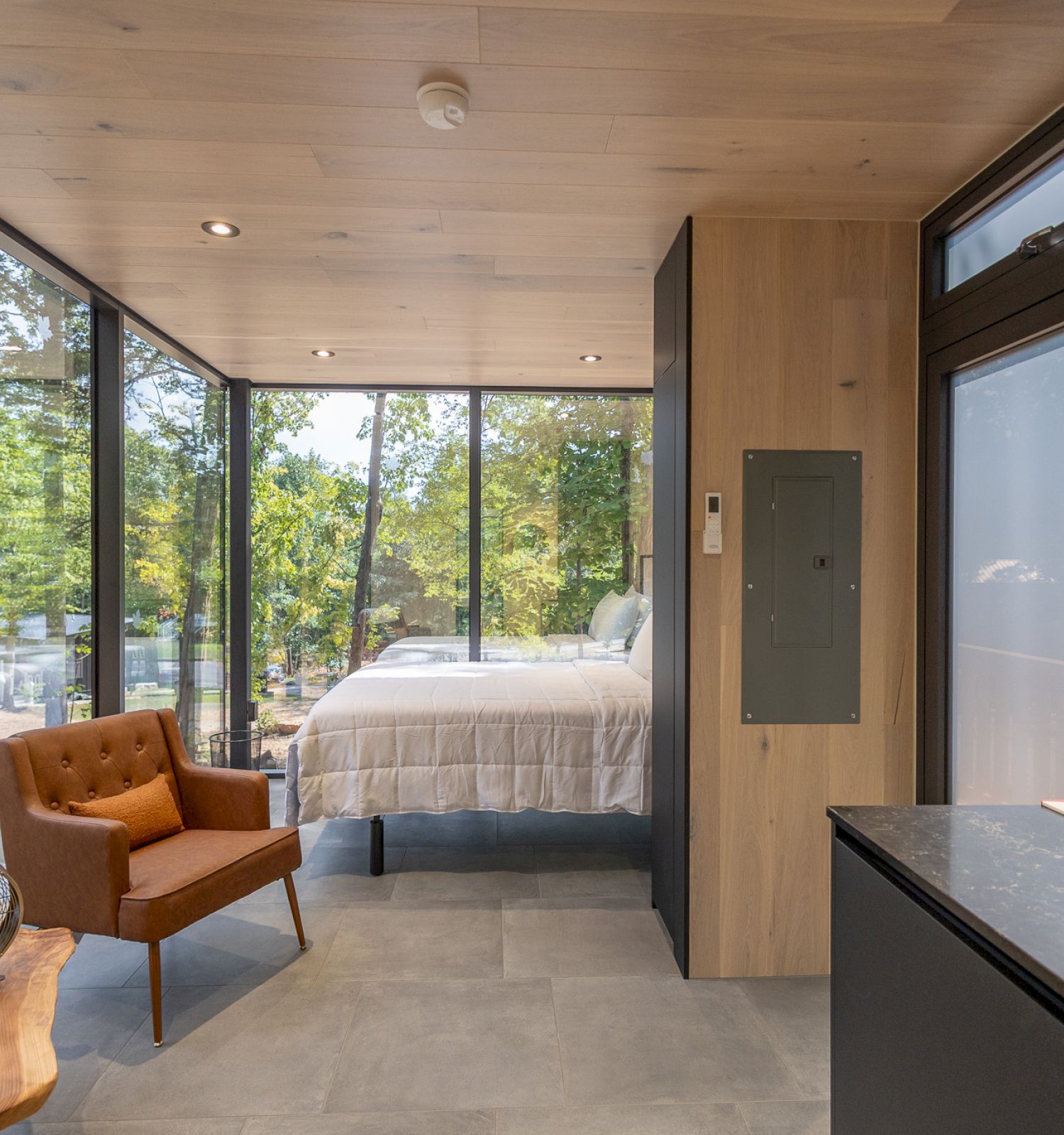 The image shows a modern interior with large windows, a bed, an armchair, and a kitchen area with an oven, surrounded by greenery outside.