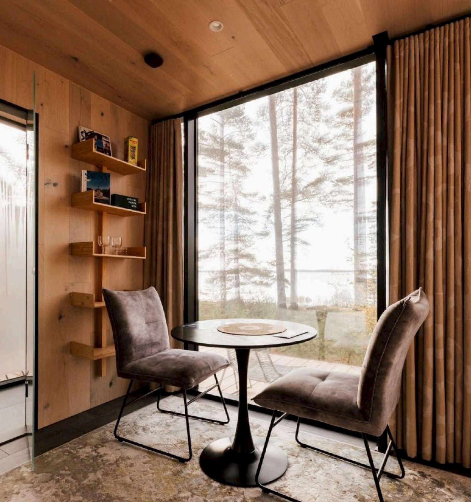 A cozy wooden interior features a round table with two chairs, a window view of trees, and a glimpse of a bathroom through an open door.