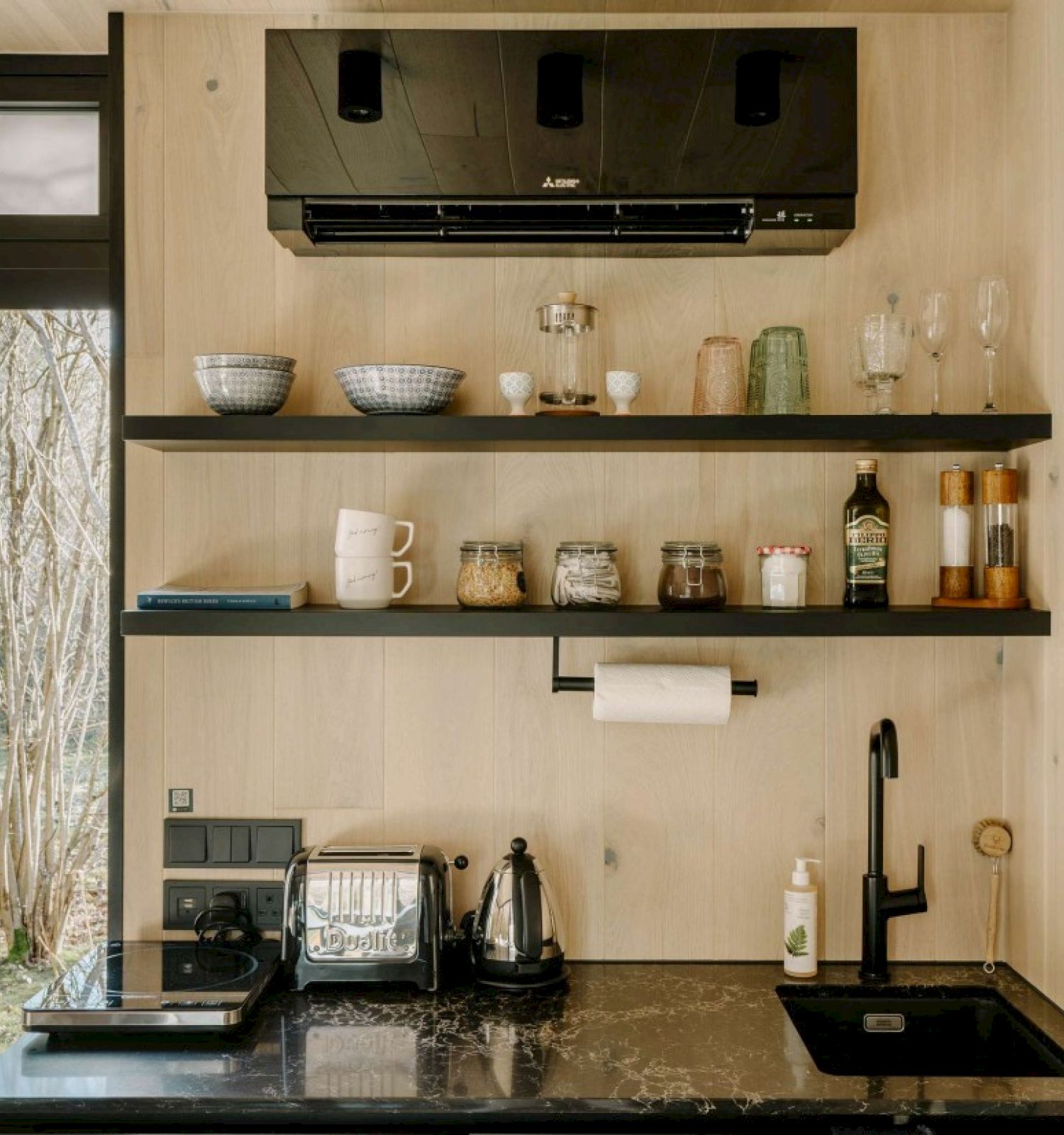 A compact kitchen area with shelves holding dishes and jars, a coffee maker, and a sink, set against a light wood wall with a window.