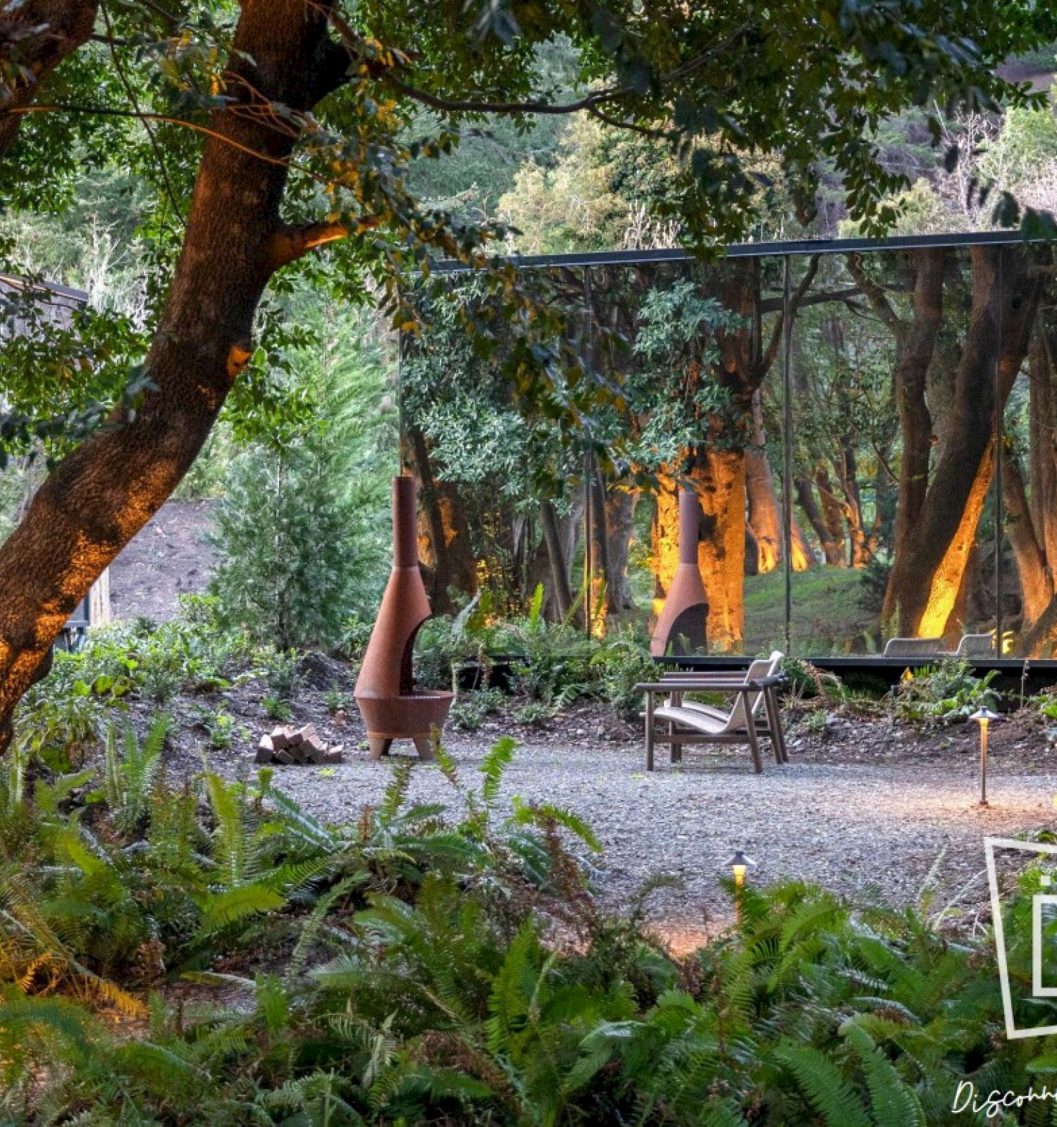 The image shows a mirrored building in a forest, reflecting trees and creating a blend with nature. A few outdoor lights illuminate the scene.