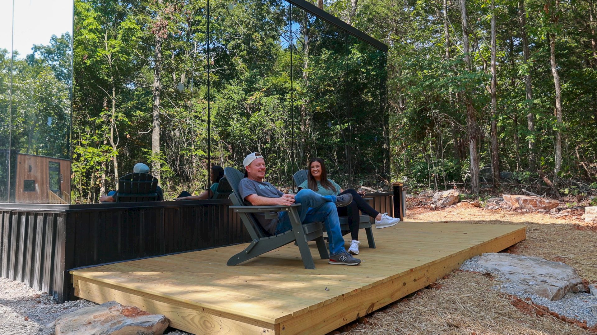 Two people are relaxing on a wooden deck in front of a modern, mirrored building surrounded by a forest setting.