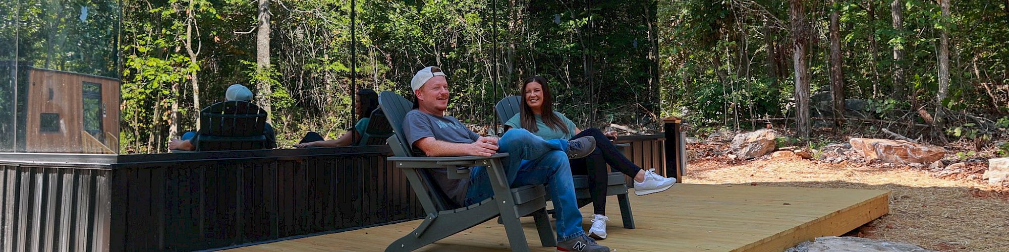 Two people sit on a wooden deck next to a mirrored building in a forested area, reflecting the surrounding trees.