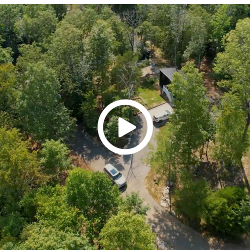 An aerial view shows a wooded area with a road, cars, and a few buildings amidst dense trees.