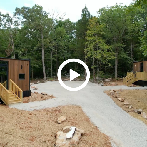 The image shows two small cabins with wooden decks surrounded by trees on a dirt path, in a forested area.