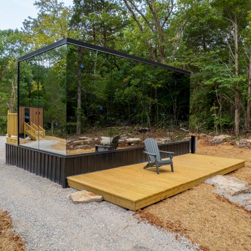 A glass structure in a wooded area with a wooden deck and a chair, surrounded by trees and gravel pathways.