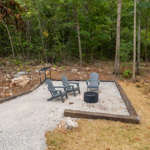 A small outdoor seating area with four chairs and a fire pit is surrounded by trees, set on a gravel patch with wooden borders.